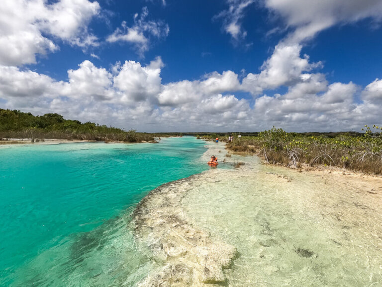 Turquoise river with greenery on the edges, against a bright blue sky with puffy clouds.