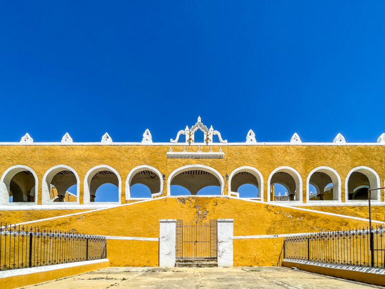 Bright yellow wall with white arches and black gates against a clear blue sky