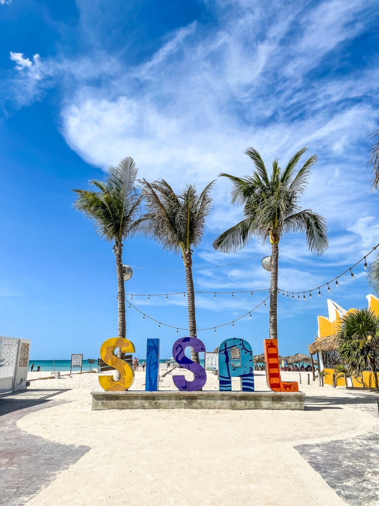 Colorful sign that says SISAL with a backdrop of palm trees and foreground of sand