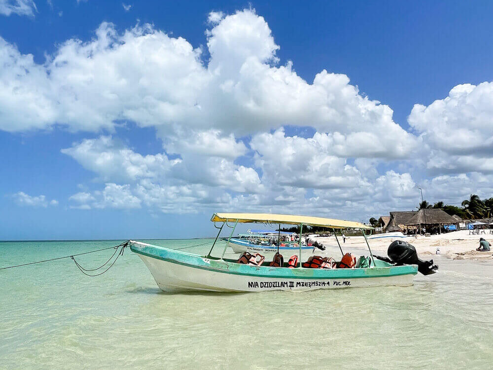 Blue and white boat floating in shallow turquoise waters against a blue sky with puffy clouds.