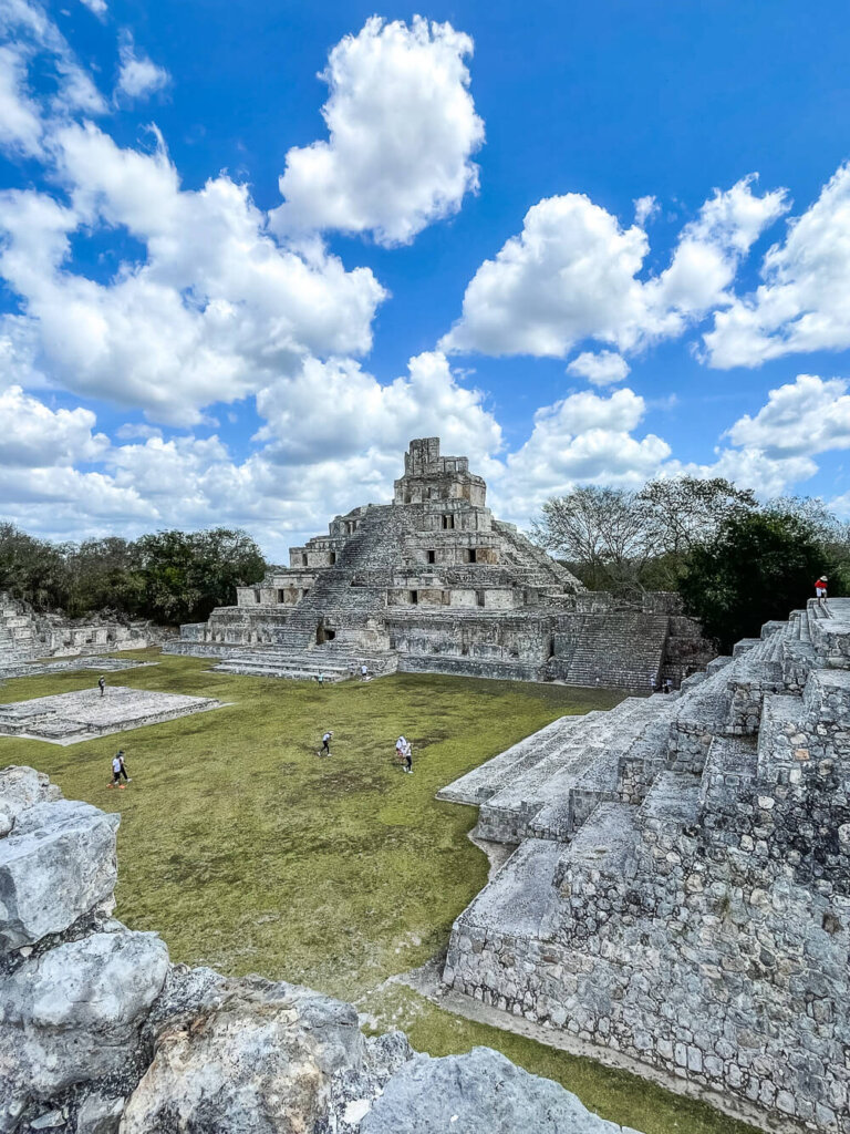 Ancient stone ruins of temples surround a grassy plaza, with a blue sky and puffy clouds