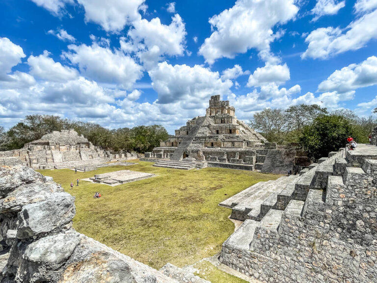 Ancient Mayan stone pyramids surrounding a grassy plaza, with blue skies and puffy clouds above