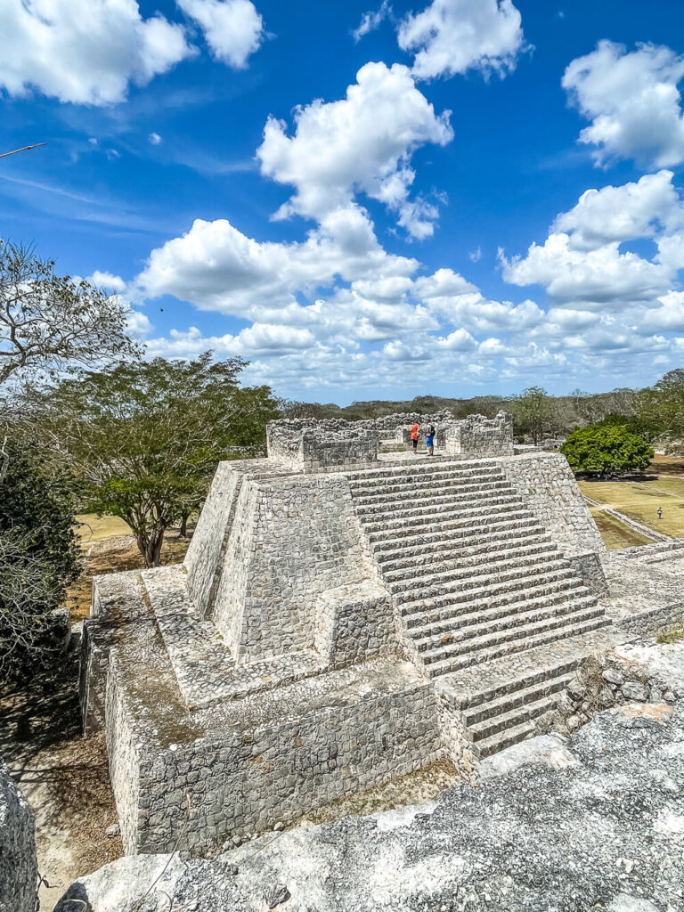 Ancient stone pyramid with people on top, against a backdrop of trees and a bright blue sky with puffy clouds.