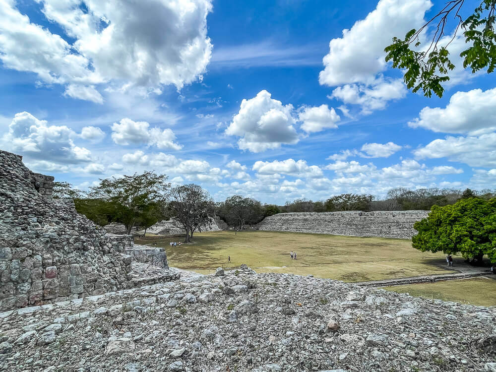 Ruins of stone temples surround a grassy plaza, with a blue sky and puffy clouds above