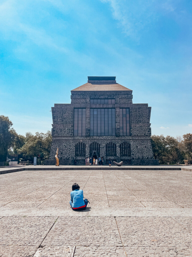 A woman in a blue shirt sits on a step of a large stone plaza, in front of a square stone building with a tower and tall windows. There are a few trees on the side of the building. The sky is blue.