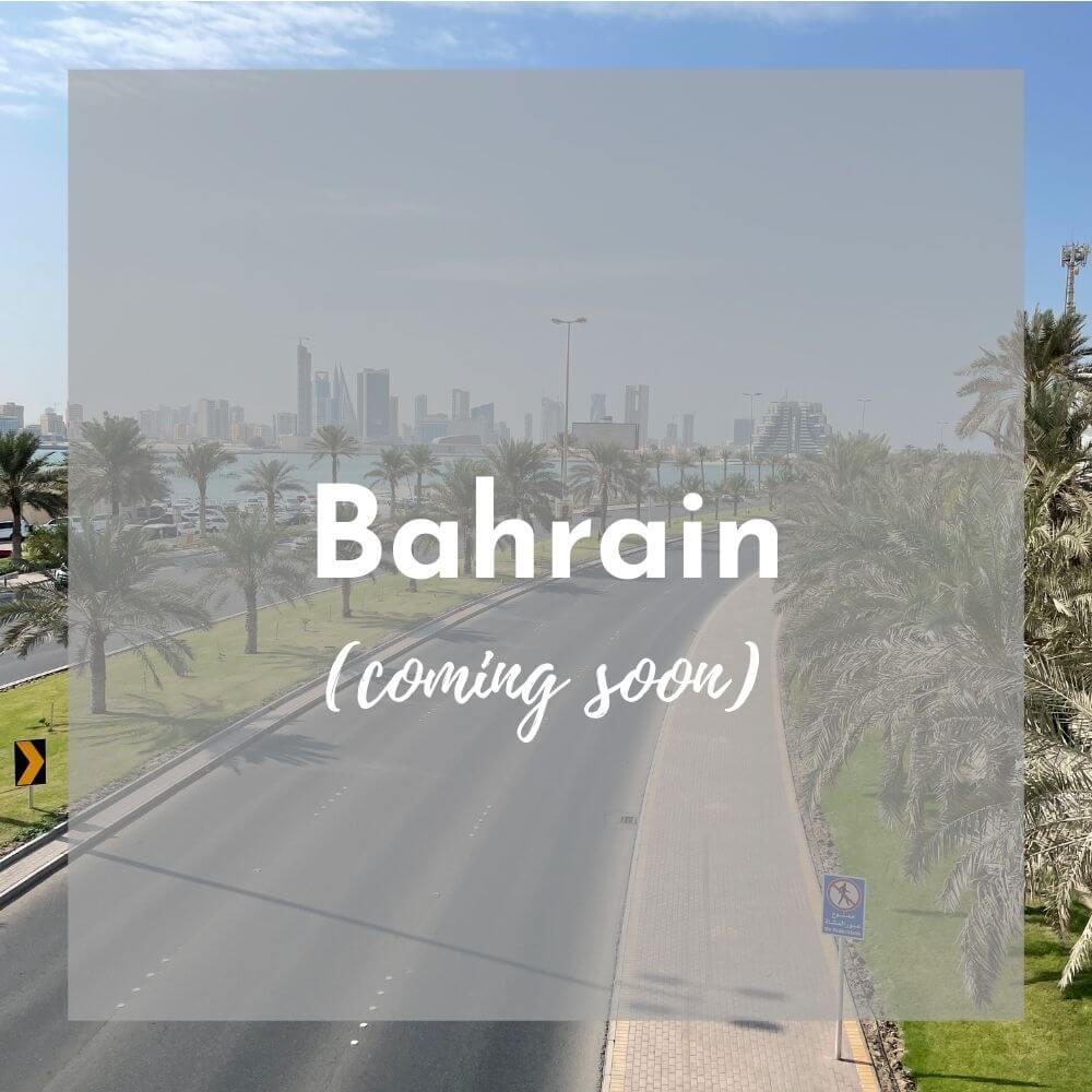 Foreground: a transparent grey box with Bahrain (coming soon) in white text Background photo: a paved road leads towards a tall skyline, with grass and trees on either side of the road, and a blue sky above