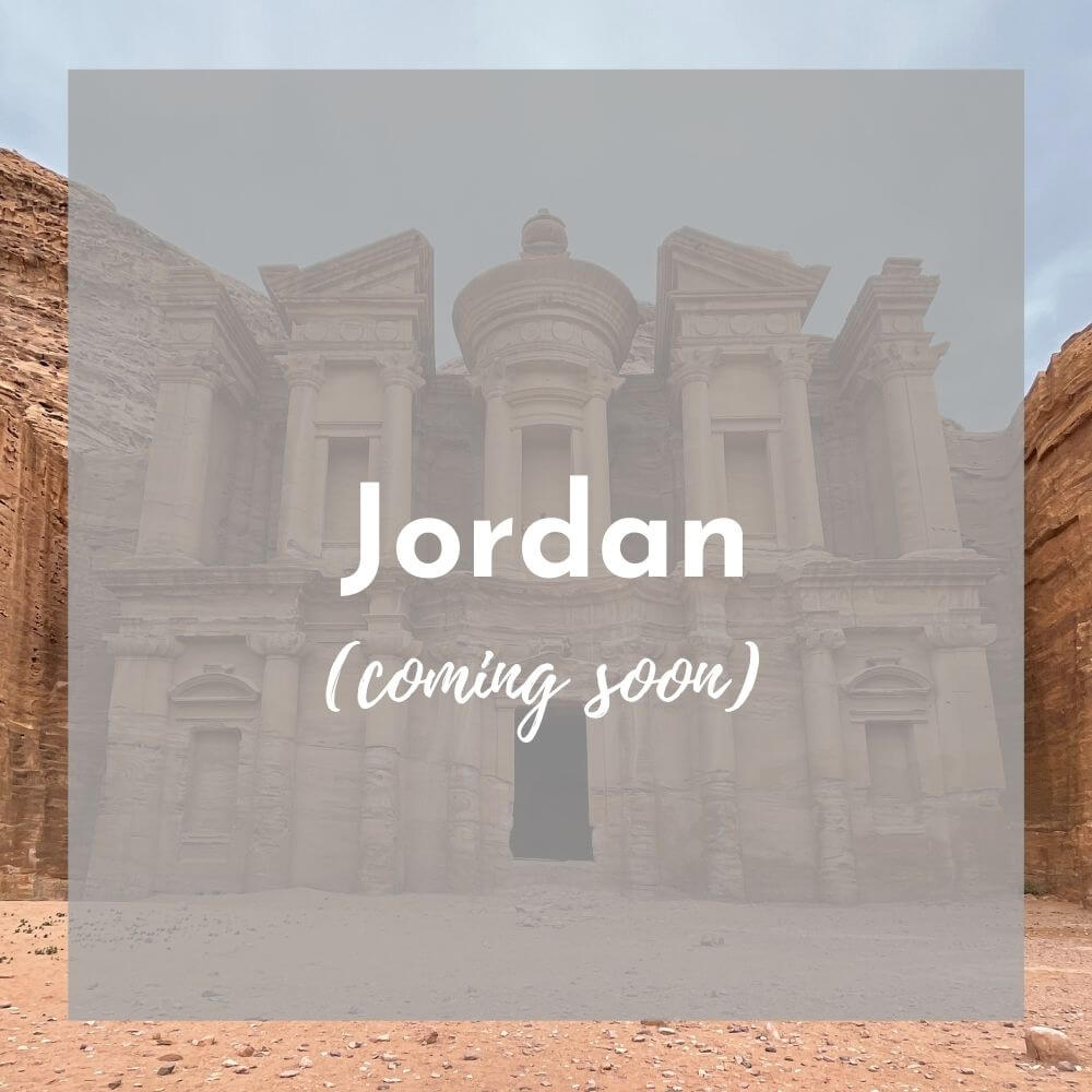 Foreground: a transparent grey box with Jordan (coming soon) in white text Background photo: a sand colored building carved out of the cliff stands tall against a blue sky