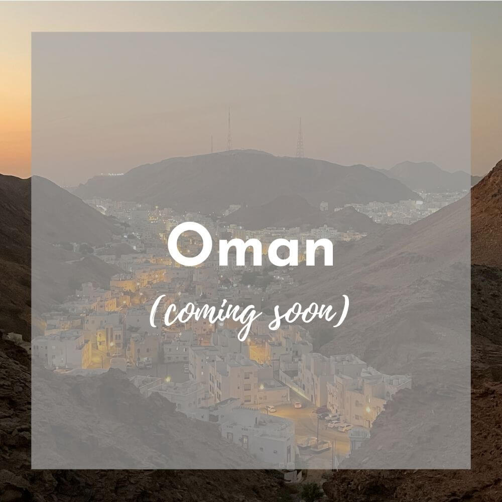 Foreground: a transparent grey box with Oman (coming soon) in white text Background photo: a sunset scene looking down on a city, with white buildings starting to glow with streetlights, nestled in the hills