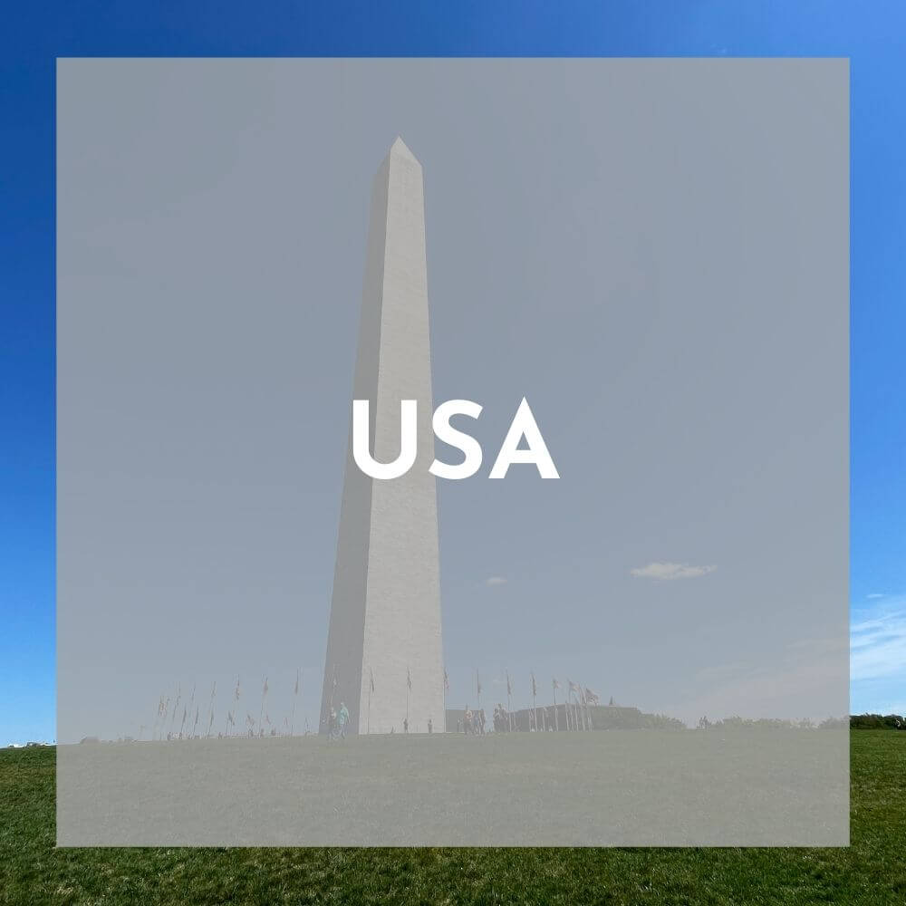 Foreground: a transparent grey box with Morocco (coming soon) in white text Background photo: a very tall marble obelisk shoots up into a blue sky. on the ground the obelisk is surrounded by flags and some people walking around