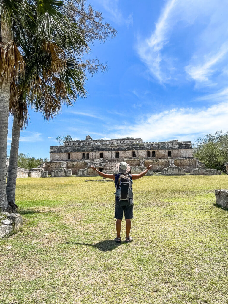 A man in a hat stands on a lawn with his arm raised. He is facing a large stone building ruin. The sky is blue with wispy clouds.