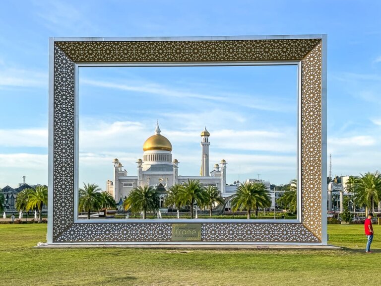 A huge frame structure with gold design on a green lawn frames a large white mosque with gold minarets in the background. A man in a red shirt stands to the right.