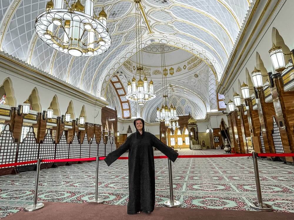 Woman in a large black cloak stands in front of a red rope inside a mosque. The walls and ceilings are white and ornately decorated.