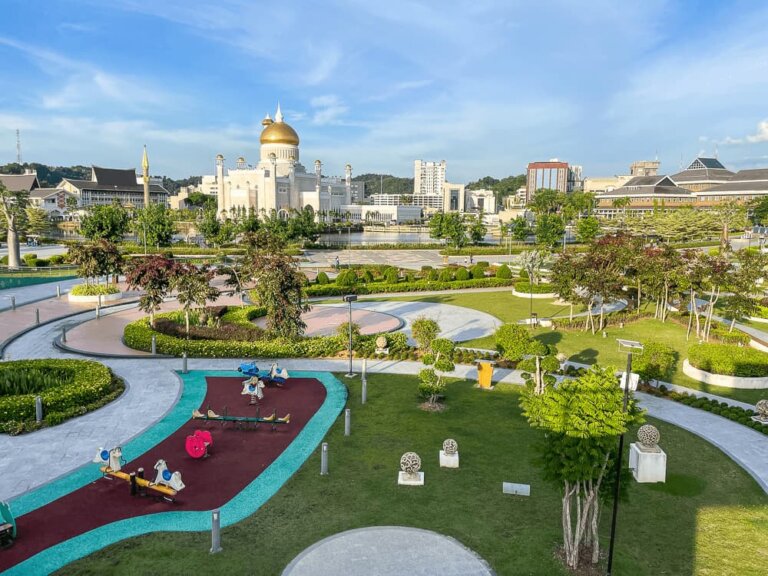 A large park with grass, bushes and playground equipment is set against the backdrop of buildings, including a large cream-colored mosque with gold domes.