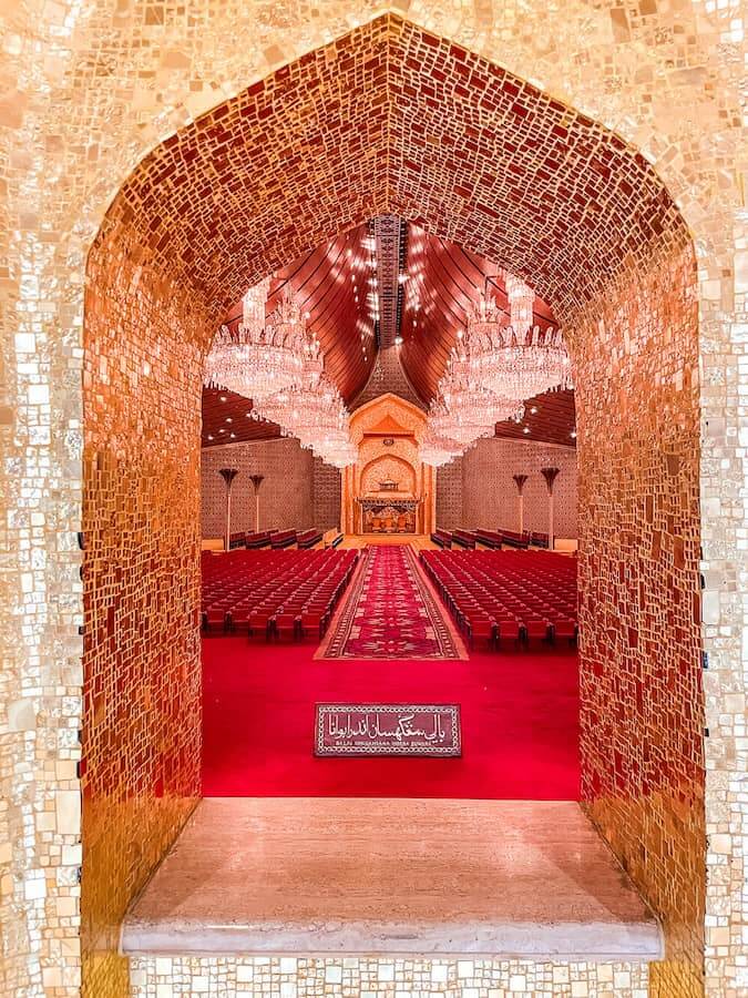 Looking through a gold-tiled window to a room with large chandeliers and red carpet and chairs neatly in a row