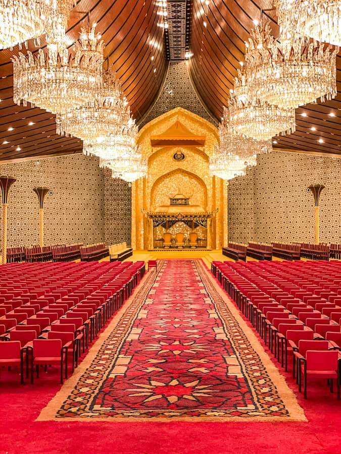 a room with large chandeliers and red carpet and chairs neatly in a row, leading up to a gold altar