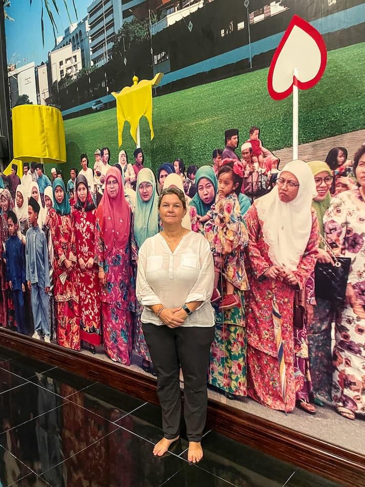 A woman in a light shirt and dark pants stands barefoot in front of a life size photo of a colorfully dressed crown of women in headscarves.