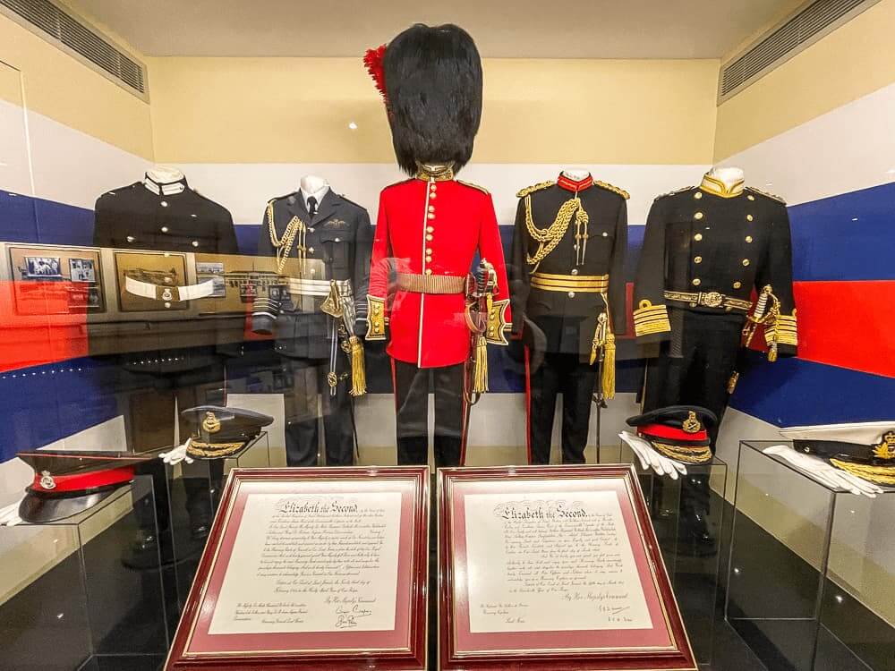 Five mannequins wearing military uniforms stand in a row. The center uniform is bright red with a large fur hat. The other uniforms are blakc with gold accents.