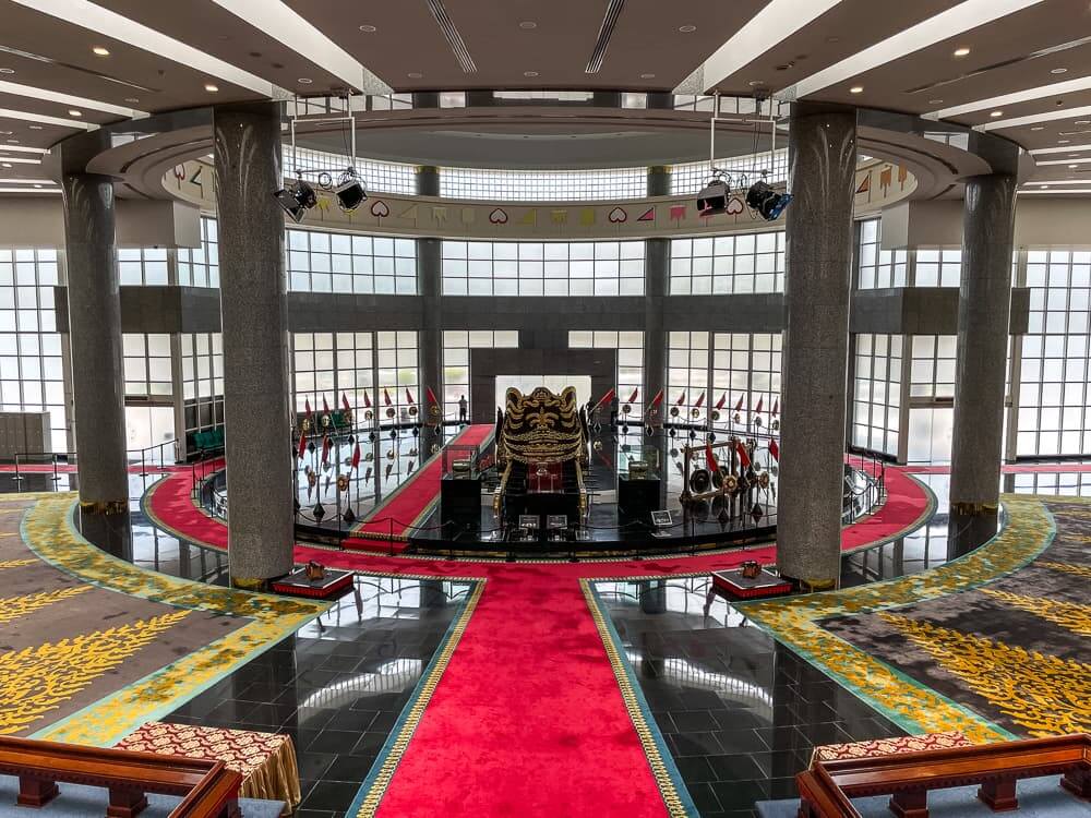 A large circular hall with large marble pillars. There is carpet on some portions of the floor. In the center is a large black and gold replica chariot.