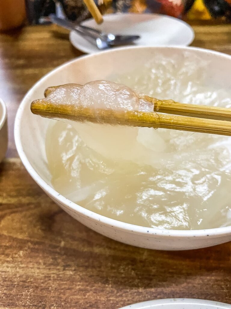 Wooden chopsticks hold up a semi-transparent gooey substance in a white bowl on a wooden table.