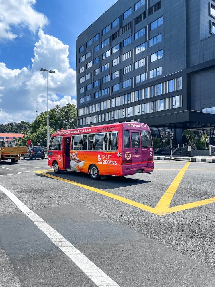 A bright pink bus is parked in a lot with yellow lines. There is a large grey building with reflective windows in the background