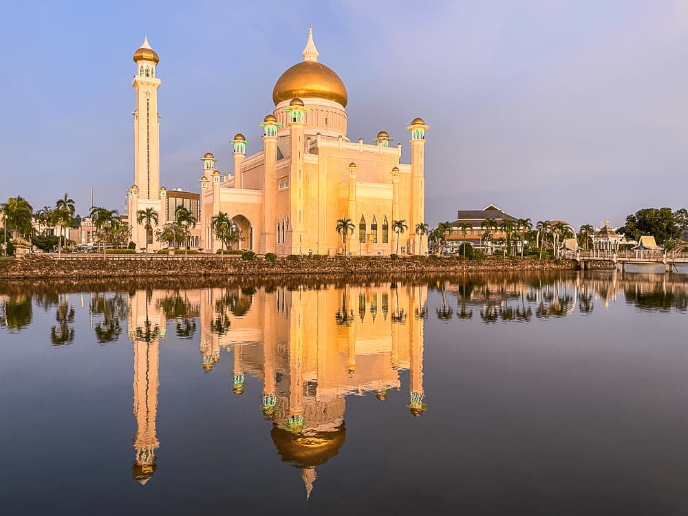 A cream-colored mosque with a golden dome and many minarets with gold domes is reflected on calm waters against a clear dusky sky