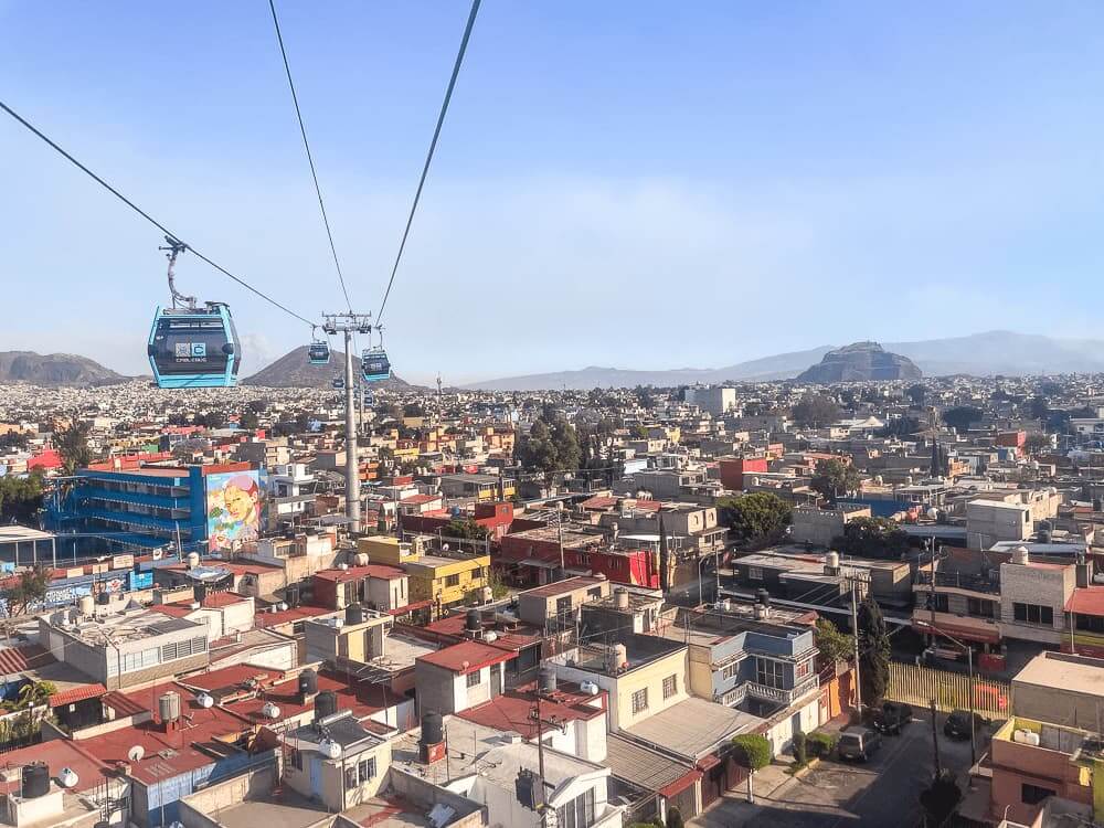 Blue cable cars float over a dense city, with mountains in the background