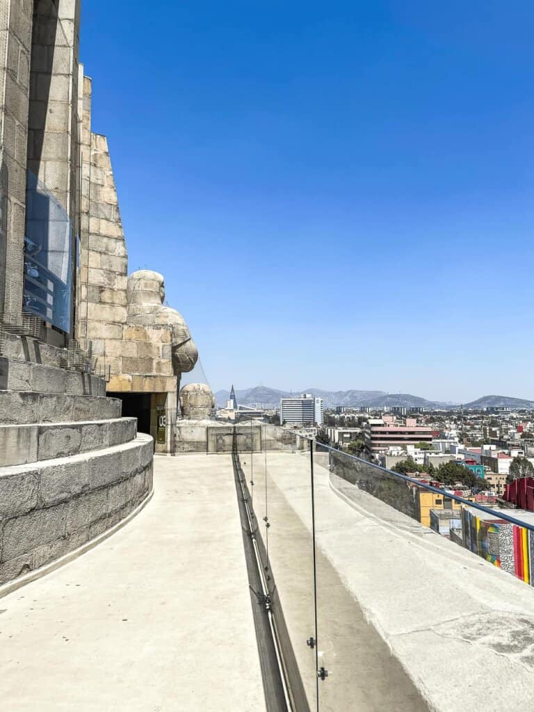 A platform with glass walls, leading to a large stone sculpture with city views in the background