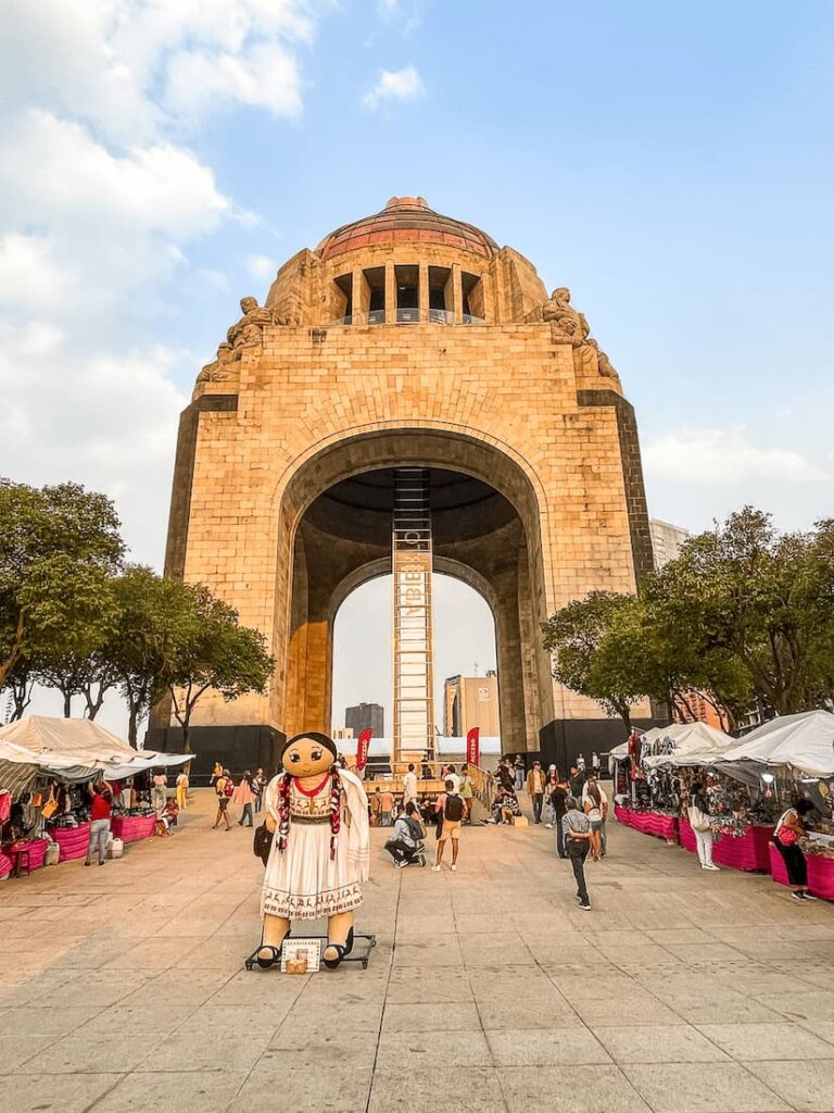 A large stone arch monument with tents leading up to it. There is a large cartoon doll in front.