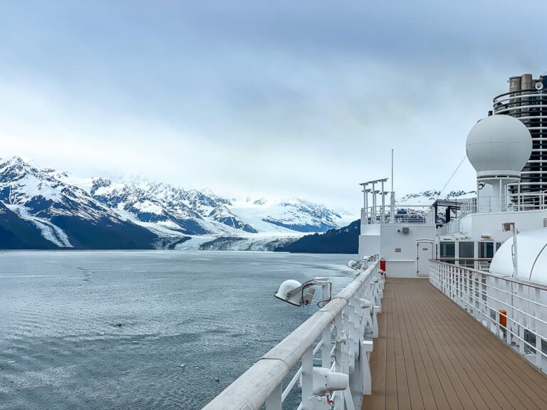 The deck of a ship on the right with dramatic snow-covered mountains in the background, against a grey sky.