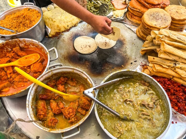 A hand reaches towards two tacos on a large grill, surrounded by steaming pots of stews.