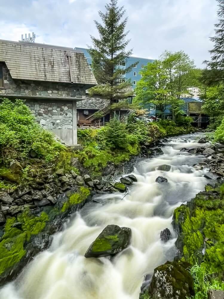 Looking up a rushing stream that runs between two grassy banks. There is a stone building on the left and a large blue building in the distance. 