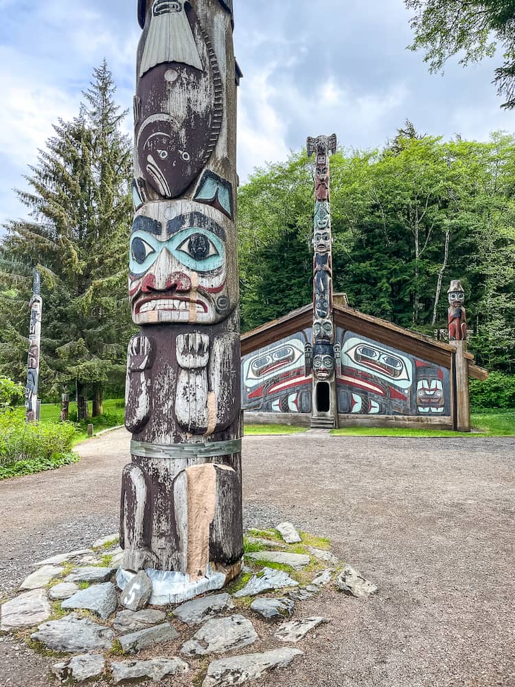 A totem pole with a red, white and blue face is in the foreground. There is a decorated wooden house in the background. 