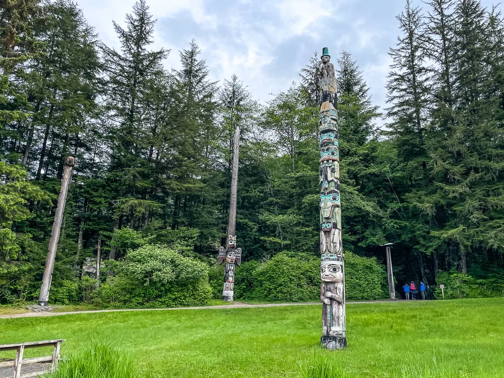 A few tall colorful totem poles stand scattered around a field, surrounded by a forest.