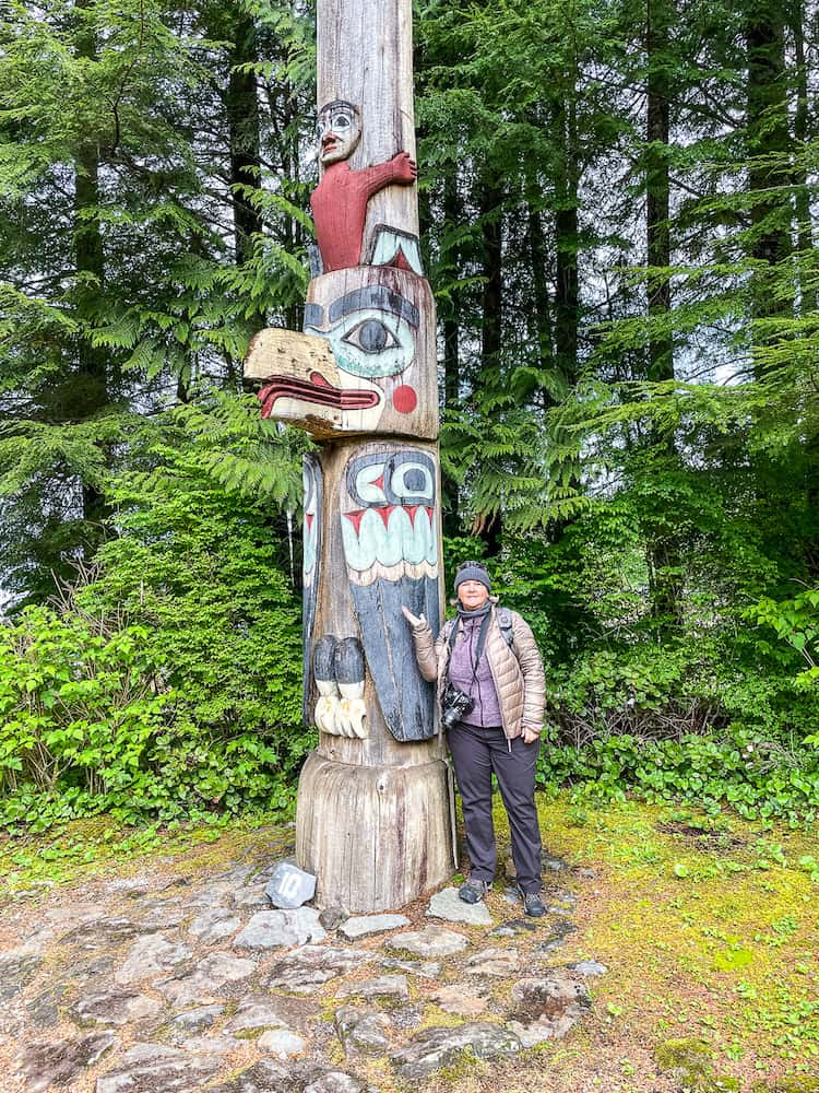 A woman in a beige jacket and grey hat leans against a tall totem pole with a bird face.