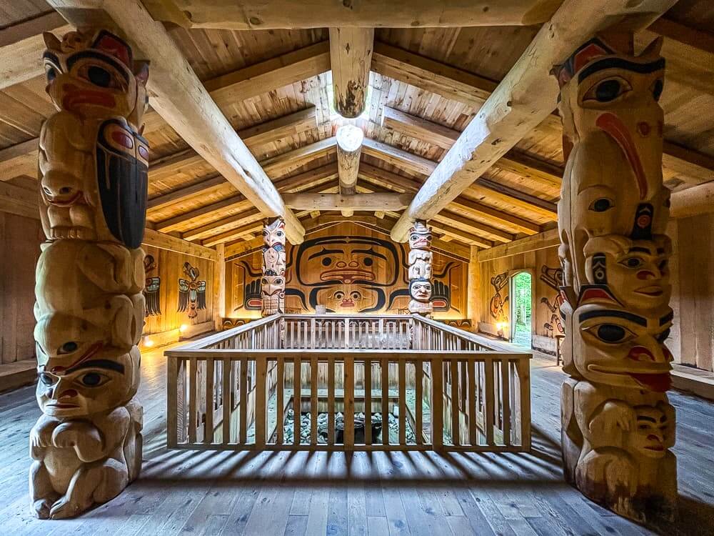 The interior of a wooden house, with log beams and decorated pillars. There is a railing in the center of the room.