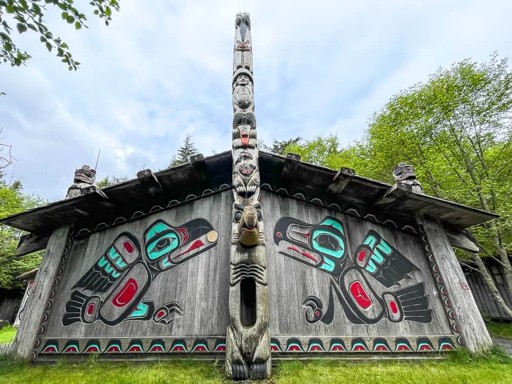 A wooden house decorated with native Alaskan paintings and a tall totem pole rising up in front.