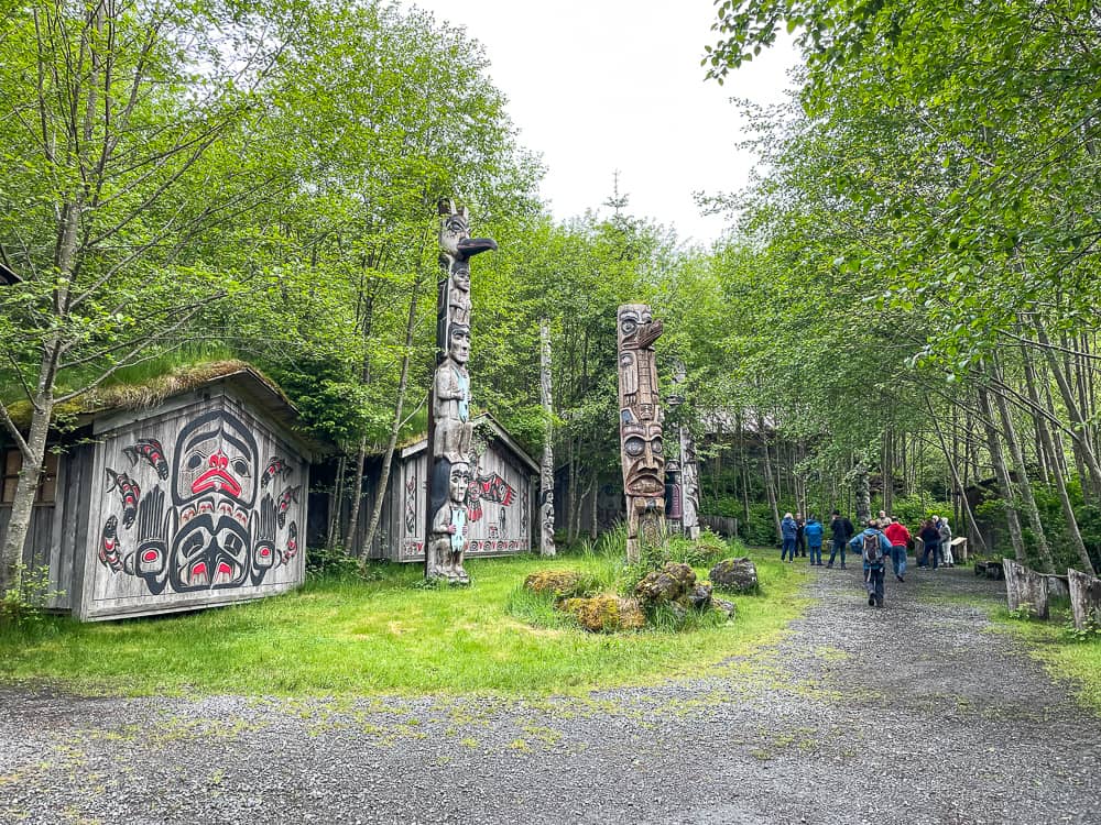 A wooden house decorated with native Alaskan paintings, set in a wooded area. Tall totem poles are scattered in front.