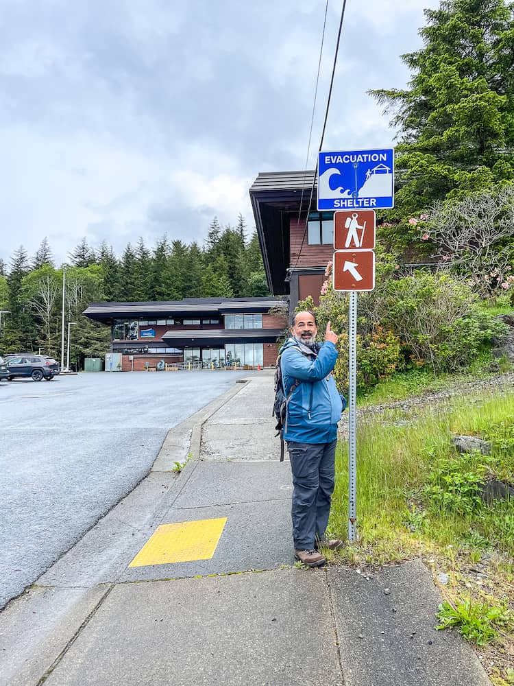 A man in a blue jacket points up at a blue and brown sign that points to a hiking trailhead.