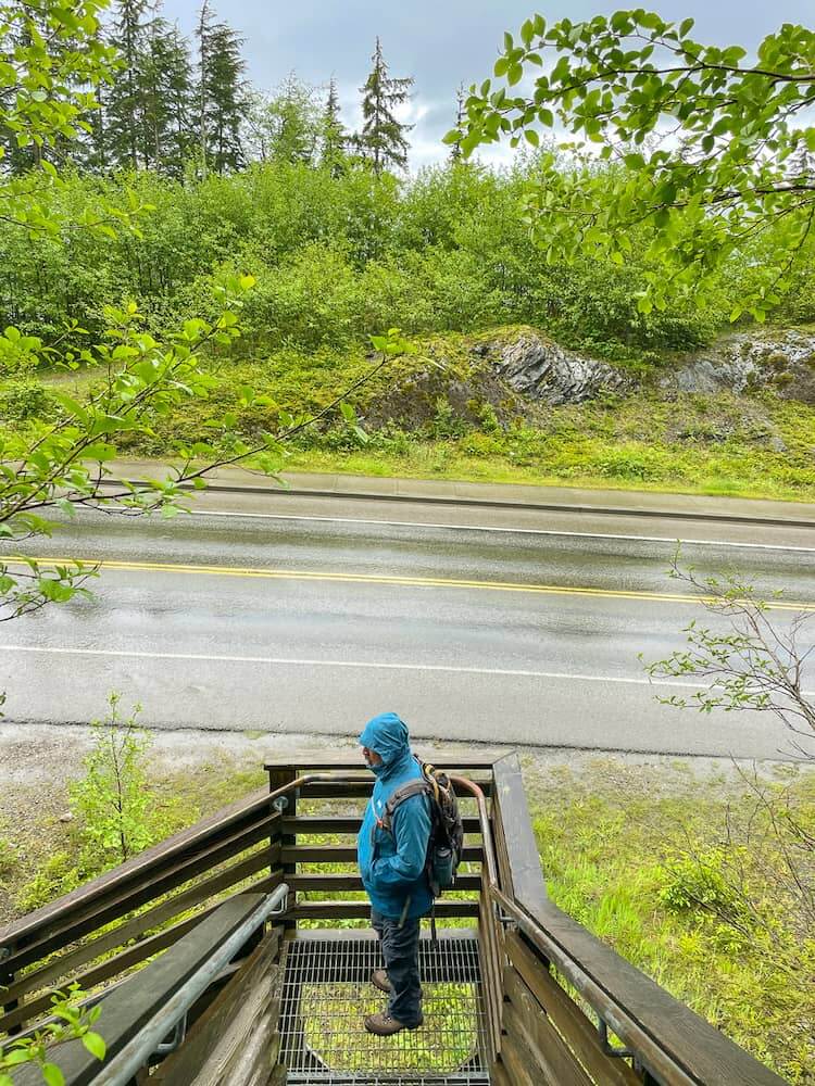 A man in a blue coat stands on a stairway leading to a road. It is raining.