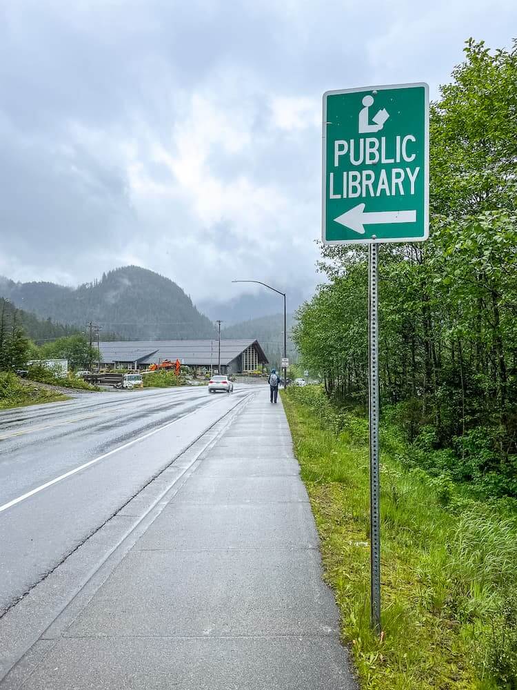 A green sign points left to the Public Library along a paved road.