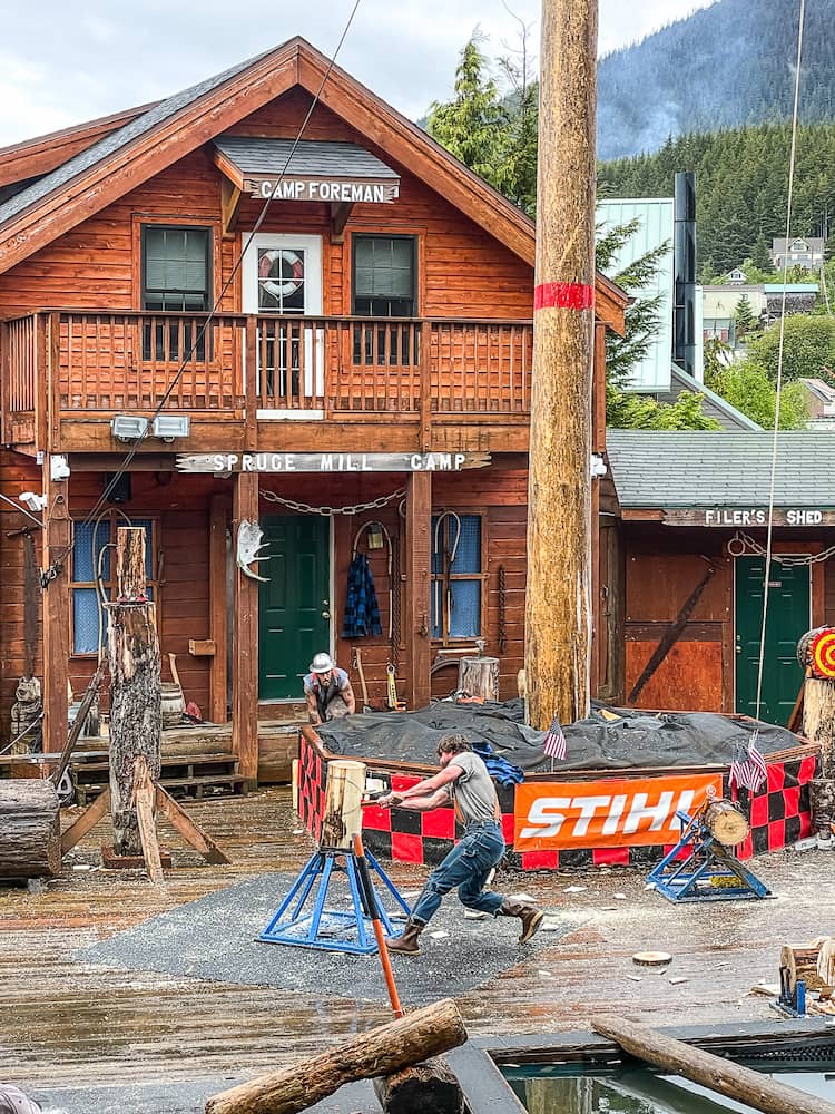 A lumberjack uses an axe to chop a stump of wood. There are wooden bulidings in the background.