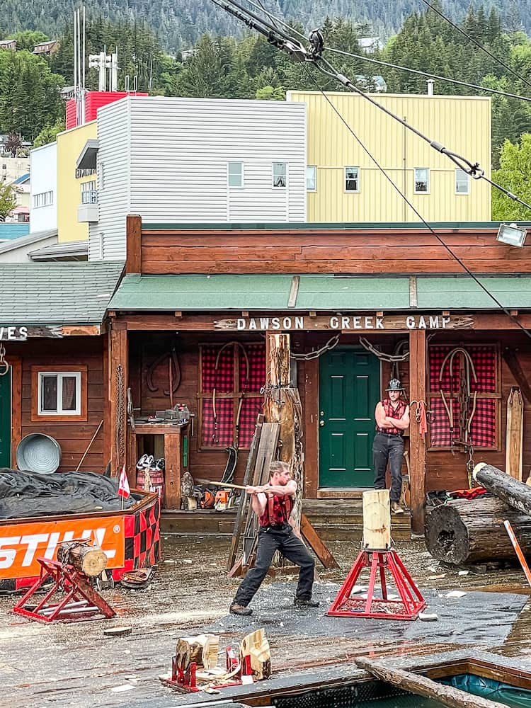 A lumberjack uses an axe to chop a stump of wood. There are wooden bulidings in the background.