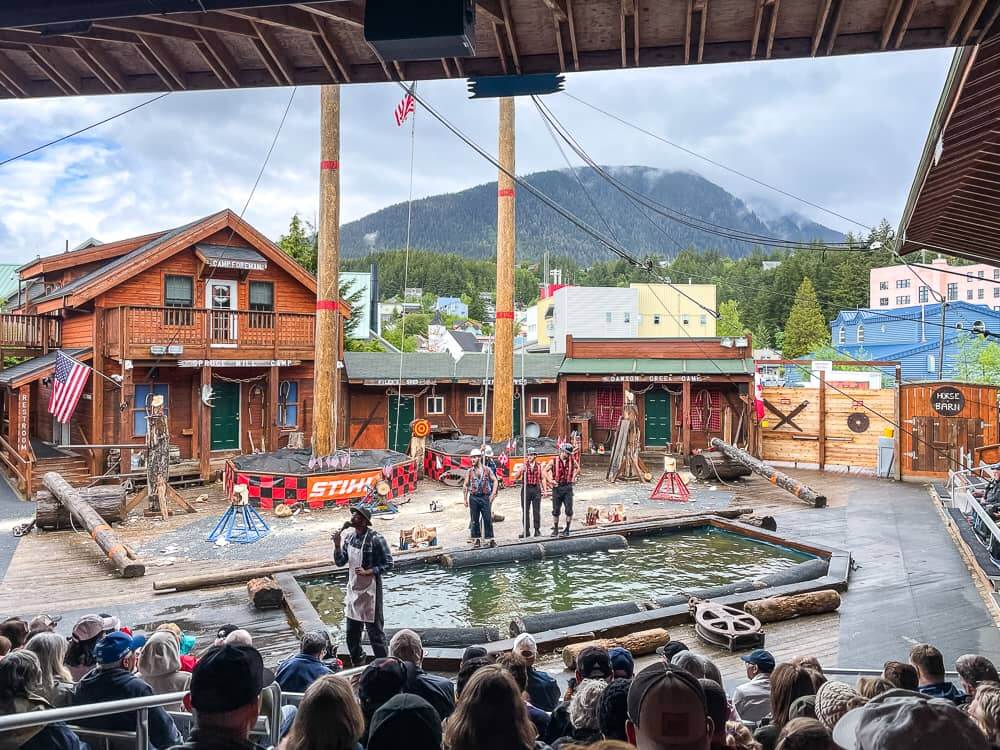 Looking down over the crowd at the Great Alaskan Lumberjack Show in Ketchikan. There is a large pool of water and two tall poles, with a series of small wooden buildings. 