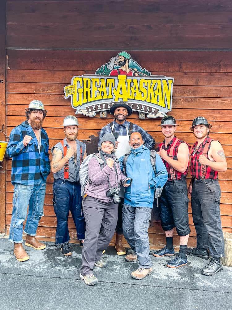 A group of lumberjacks dressed in plaid, and two tourists pose for a photo under a yellow sign.