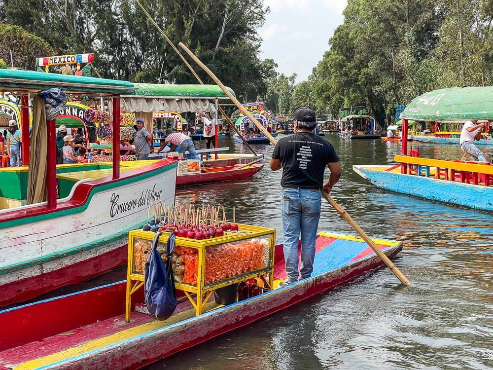 A man stands on a boat, using a long pole to guide it. There is a case of candied apples on the boat.