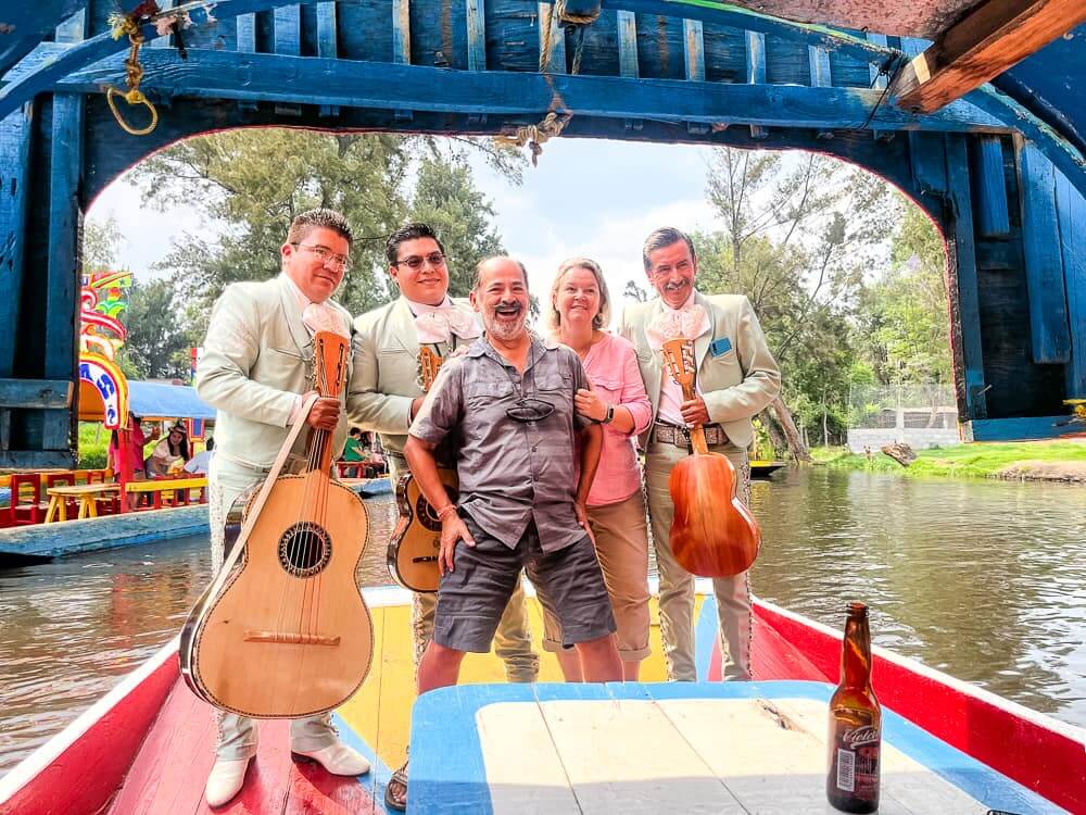 Five people stand at the back of a flat-bottomed boat. Three of the men are Mexican mariachis dressed in nice light green suits. They hold their instruments - a large guitar and small guitar.