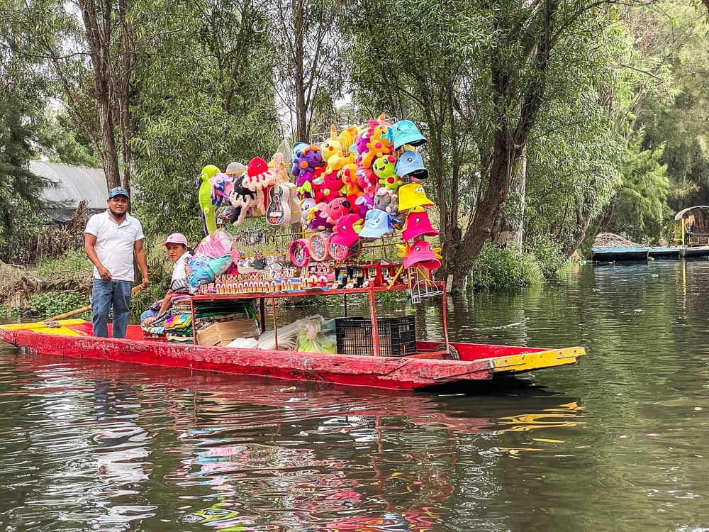 A red and yellow flat-bottomed boat floats down a canal. There are stacks of colorful souvenirs on board.