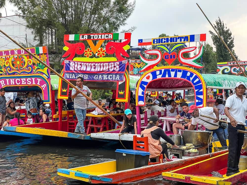 Colorful flat-bottomed boats clustered together on a small canal. A man with a pole stands on the back of one of the boats.