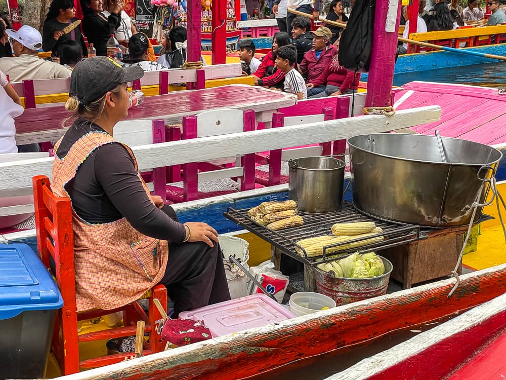 A woman in a red apron sits on a boat with corn on the cob laid on grill in front of her.