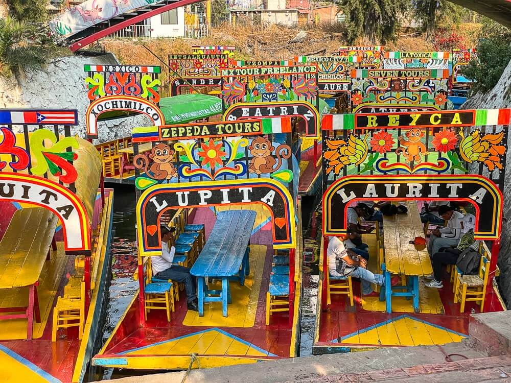 A bunch of colorful boats are parked up next to each other. They all have entrance archways with names painted on them, topped with decorative flower designs.